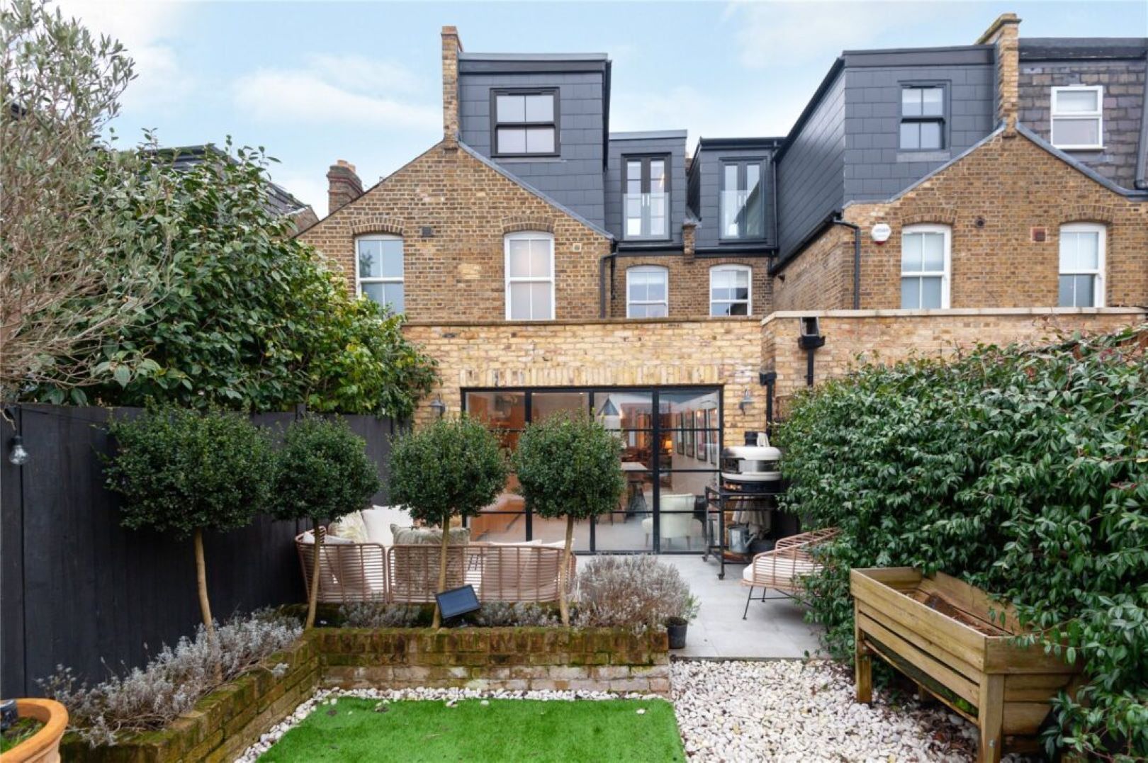 A London garden with seating area at the back and grassy area at the front, separated by topiary trees.
