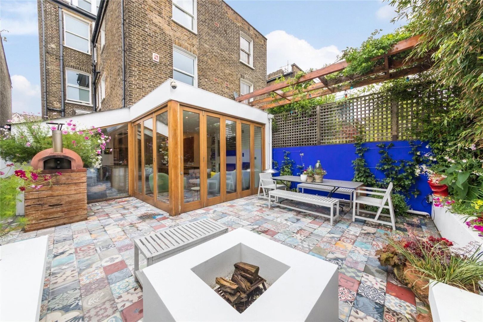 A London garden with large doors leading into the house. The garden has colourful patterned tiles and has a pizza oven t othe left and a fire pit.