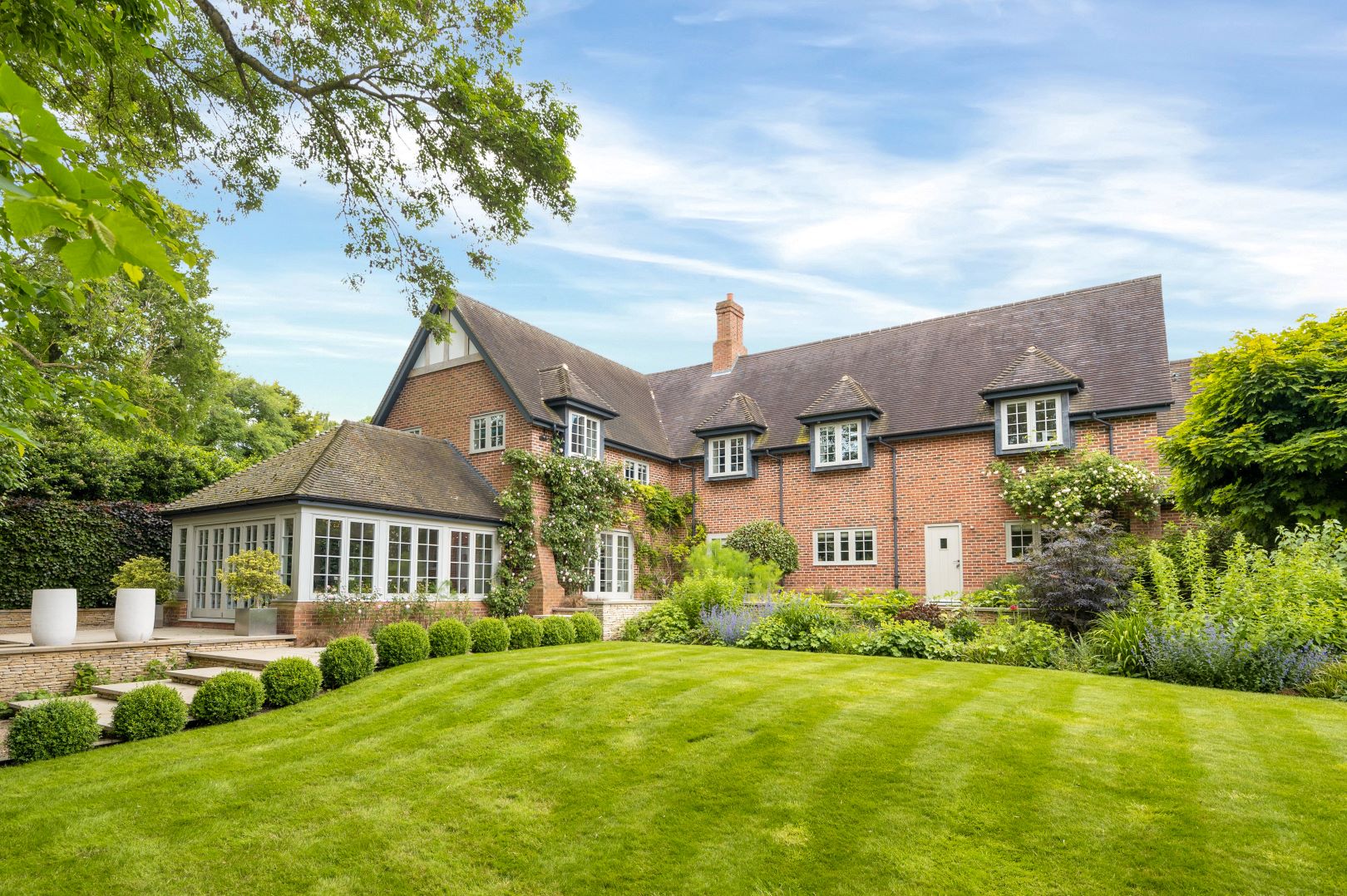 Red brick house with slate roof sitting behind a mown lawn bordered with flower beds and round topiary balls. 
