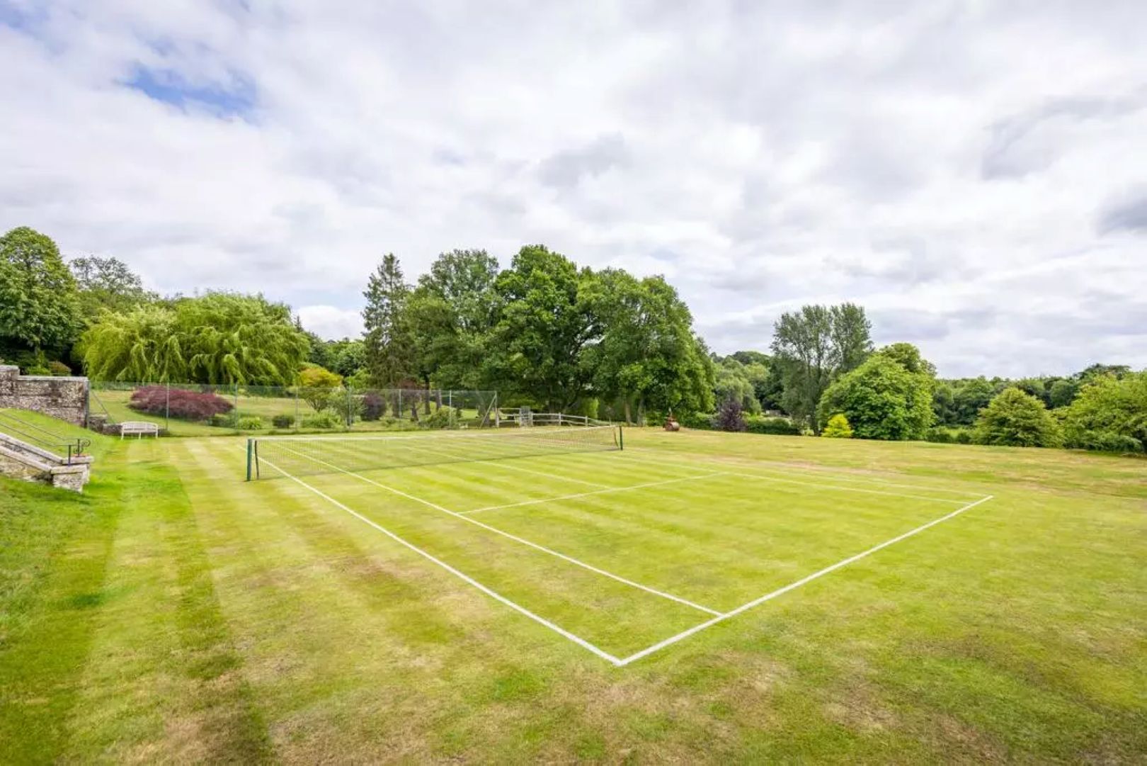 Grass tennis court with trees in the background and stone staircase to the left.