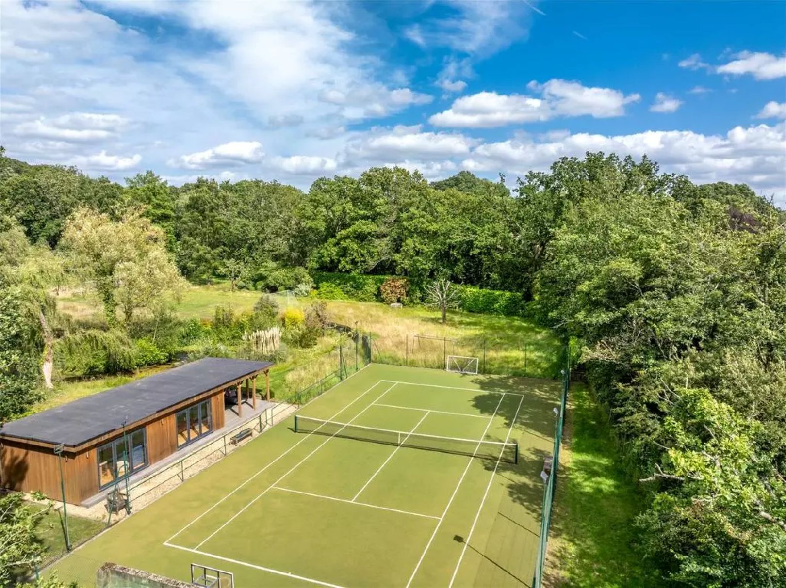 A grass tennis court with wooden pavilion to the left hand side and trees around the complex. 