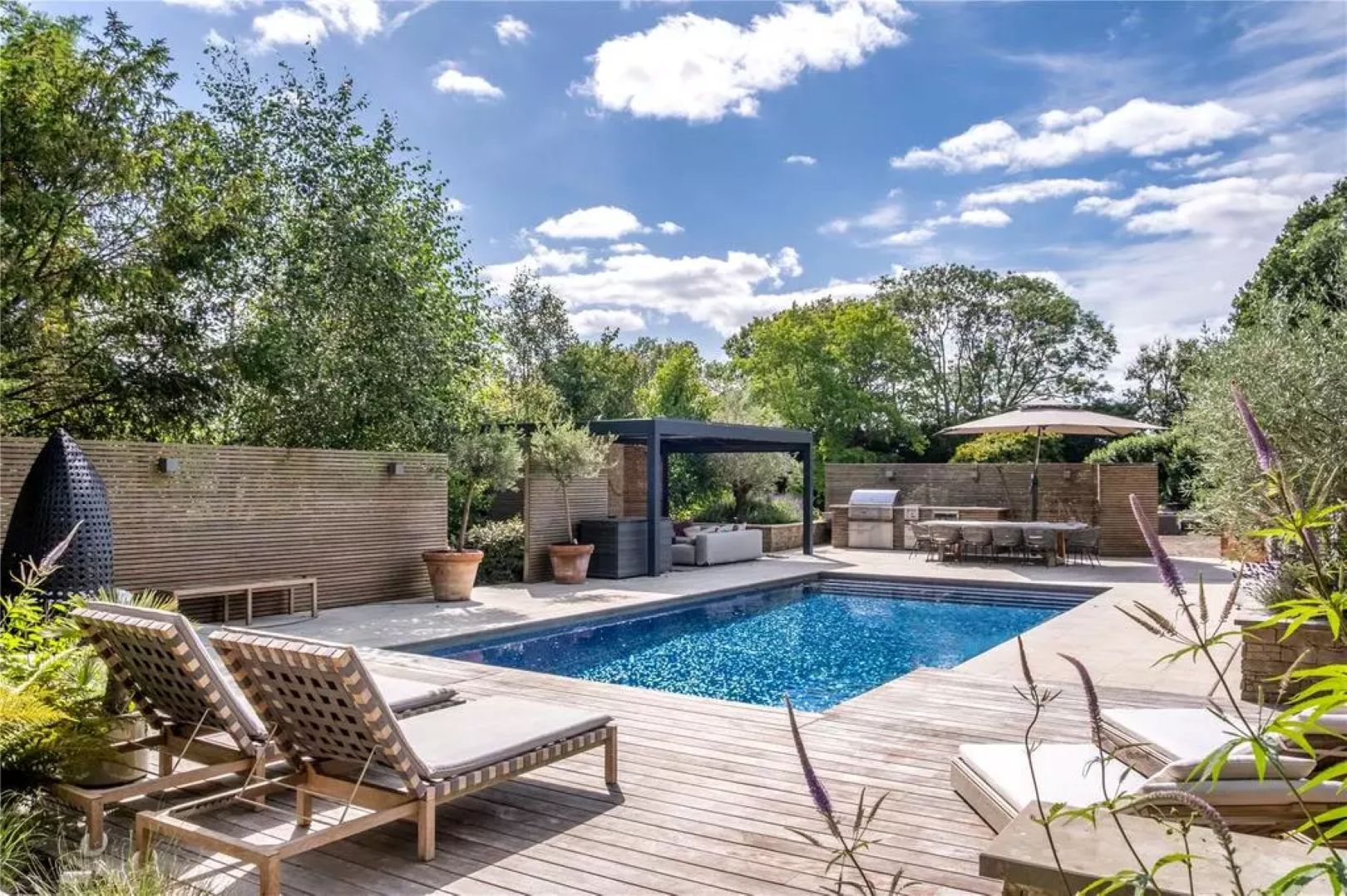 A swimming pool surrounded by a wooden terrace, with sun loungers to the front and a outdoor kitchen and dining area to the rear
