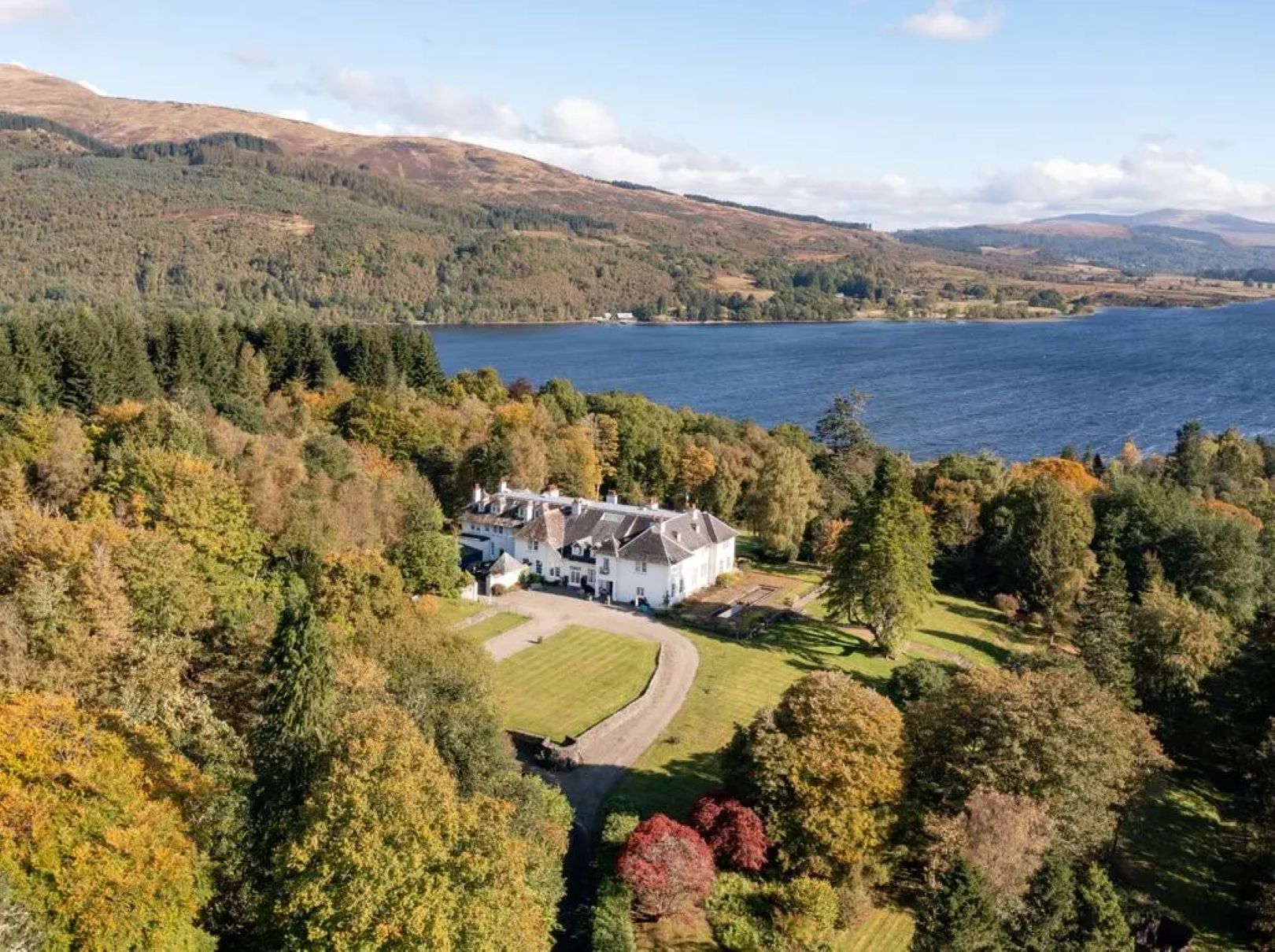 A white house sitting among woodland with a lawn and driveway to one side and a loch in the background.
