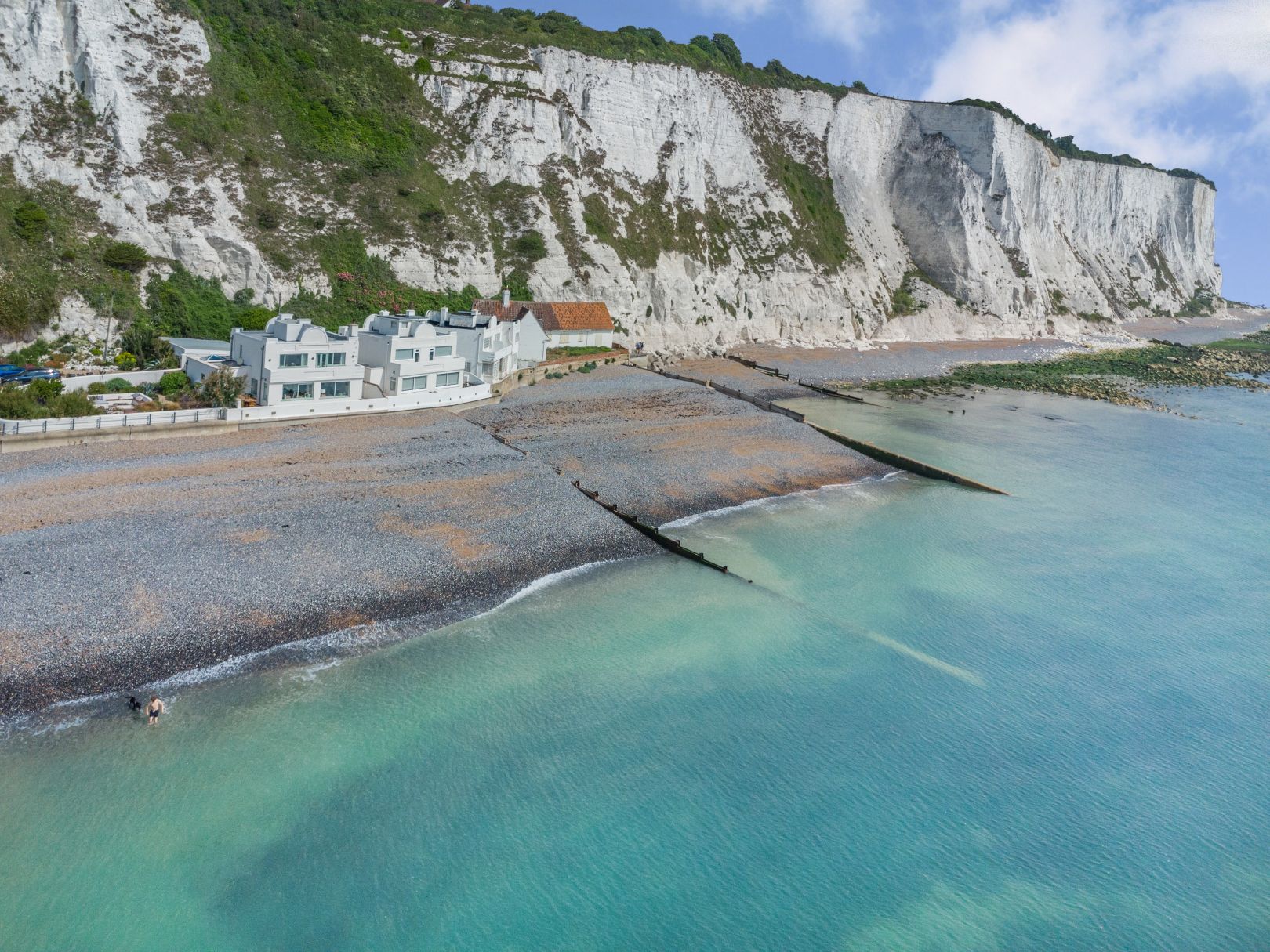 White Cliffs of Dover with a few white houses in front of them. A pebble beach and turquoise sea sits in front. 