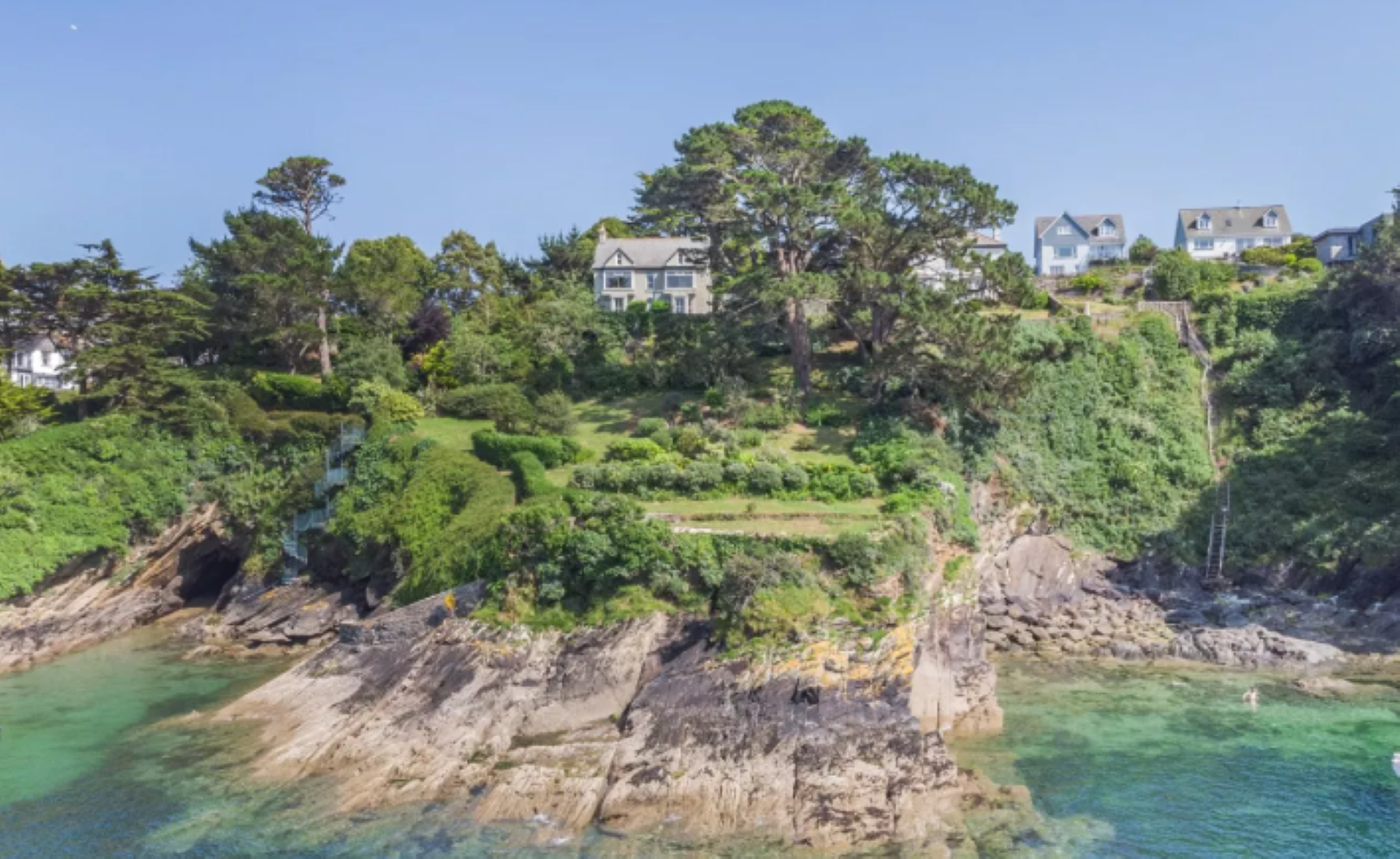 A grey house at the top of a hill, above a cliff which falls into turquoise water. 