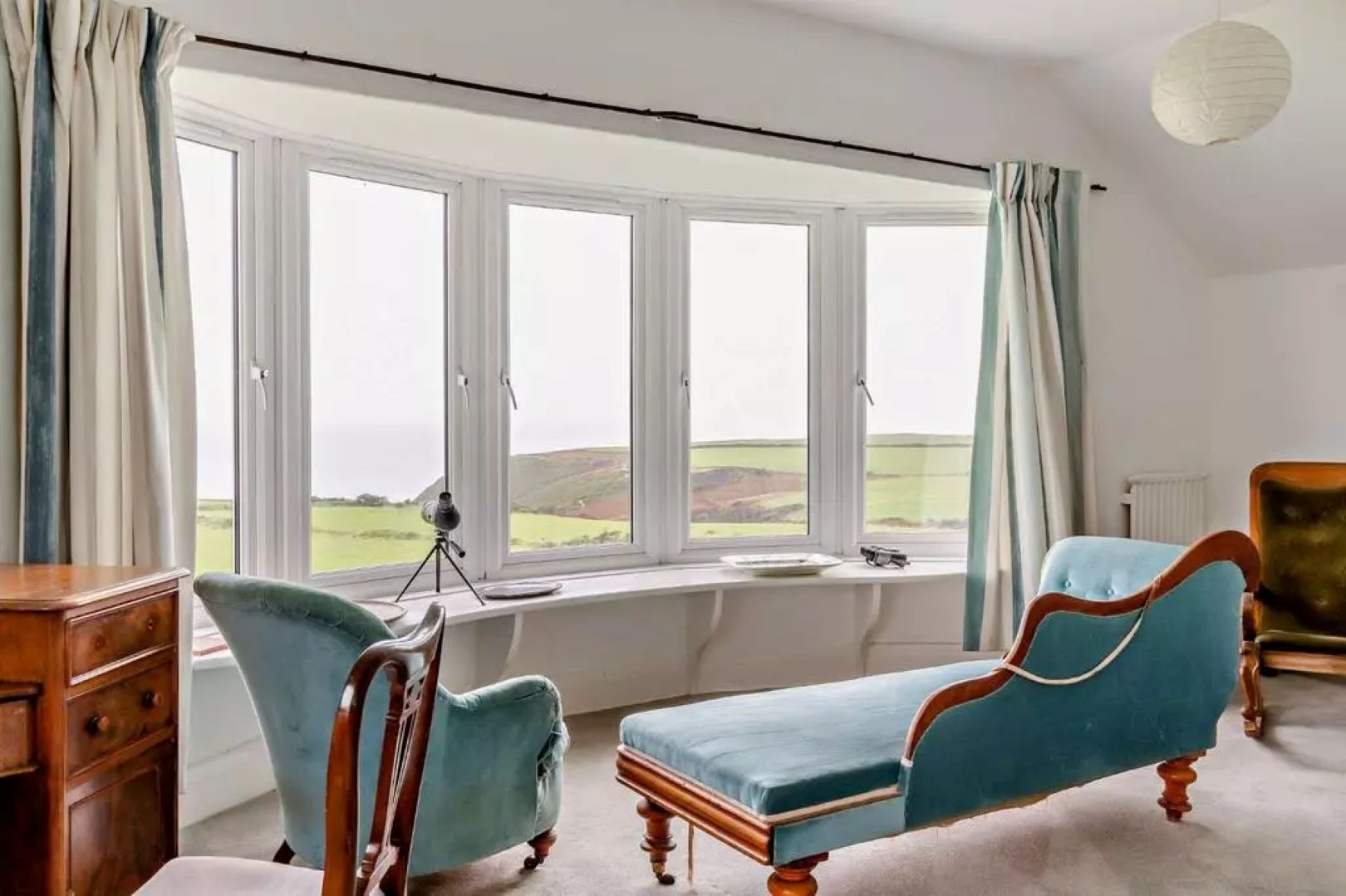 A white room with a blue chaise longue looking out of a panoramic window, looking over cliffs.