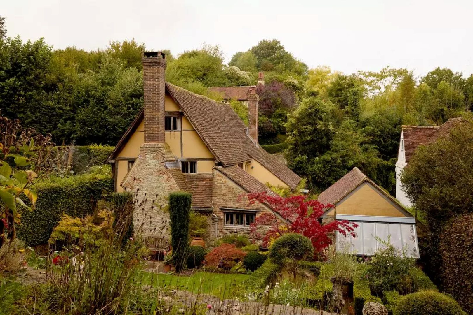 A yellow and stone old house with tiled roof sitting among shrubs and trees