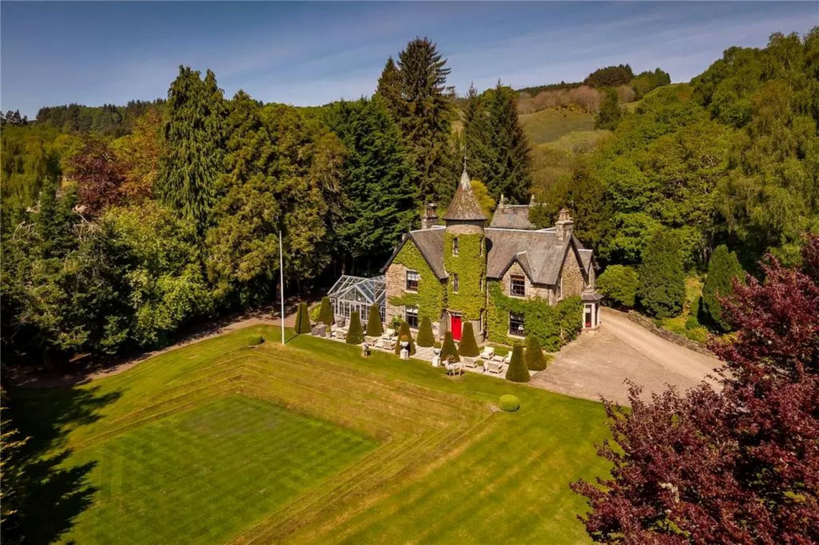 A house with ivy growing up the side and a turret at the front, with a bright red door. A lawn sits at the front with woodland at the back. 
