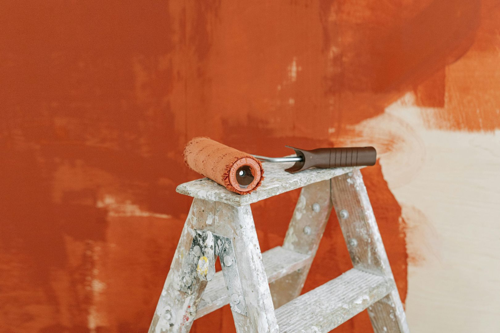 A half painted wall, from white to orange, with a step ladder in front and paint roller on top