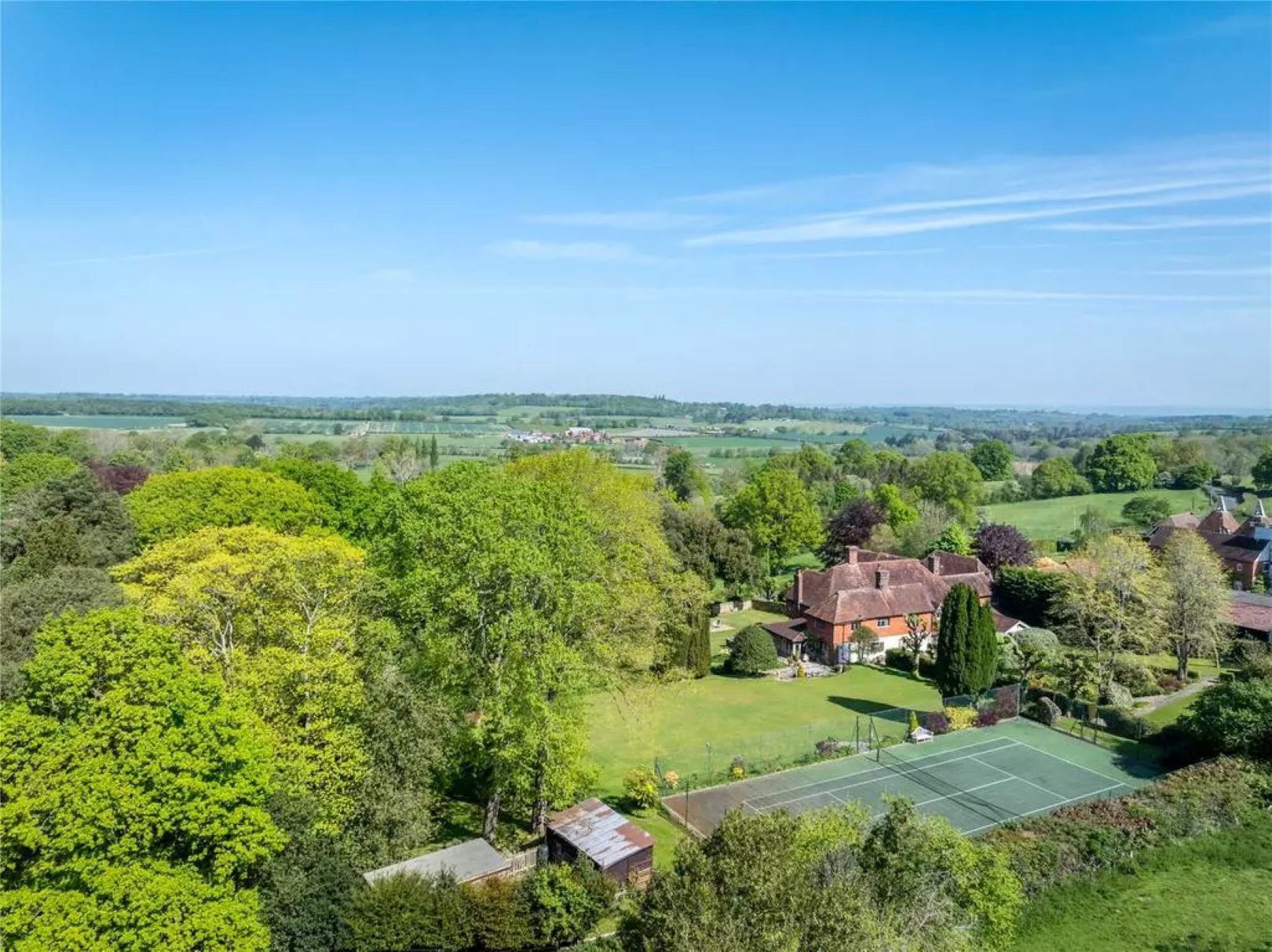 An aerial views of a white and red brick house, with a large garden, lots of trees and a tennis court
