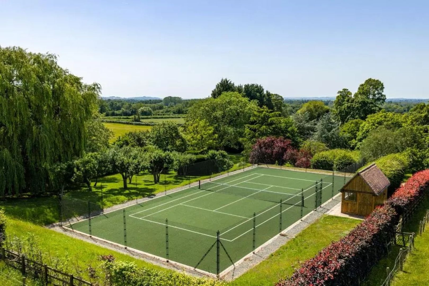 A hard tennis court with hedges and a wooden summer house