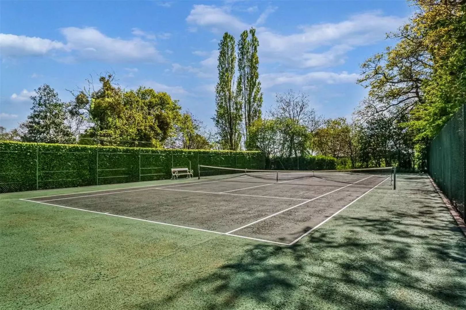 A hard tennis court with white bench overlooking the net, the court is surrounded by green hedges
